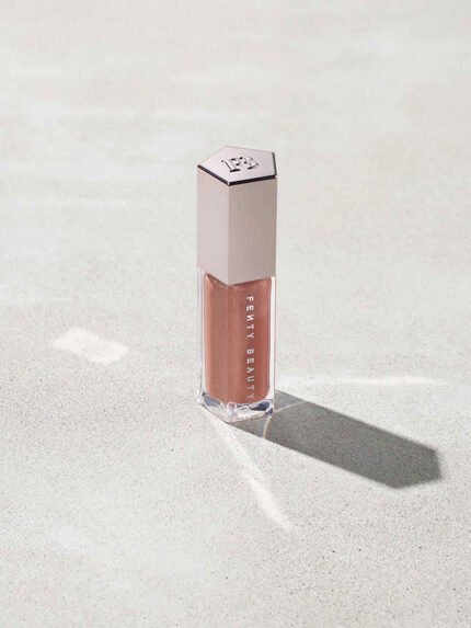 A closed tube of Gloss Bomb Universal Lip Luminizer in the shade Fenty Glow on a grey background.