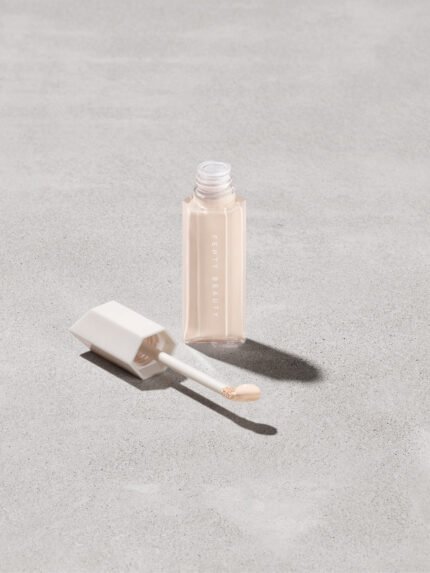 Open bottle of Fenty Beauty We're Even Hydrating Longwear Concealer with doe foot applicator in shade 100c on a concrete background.