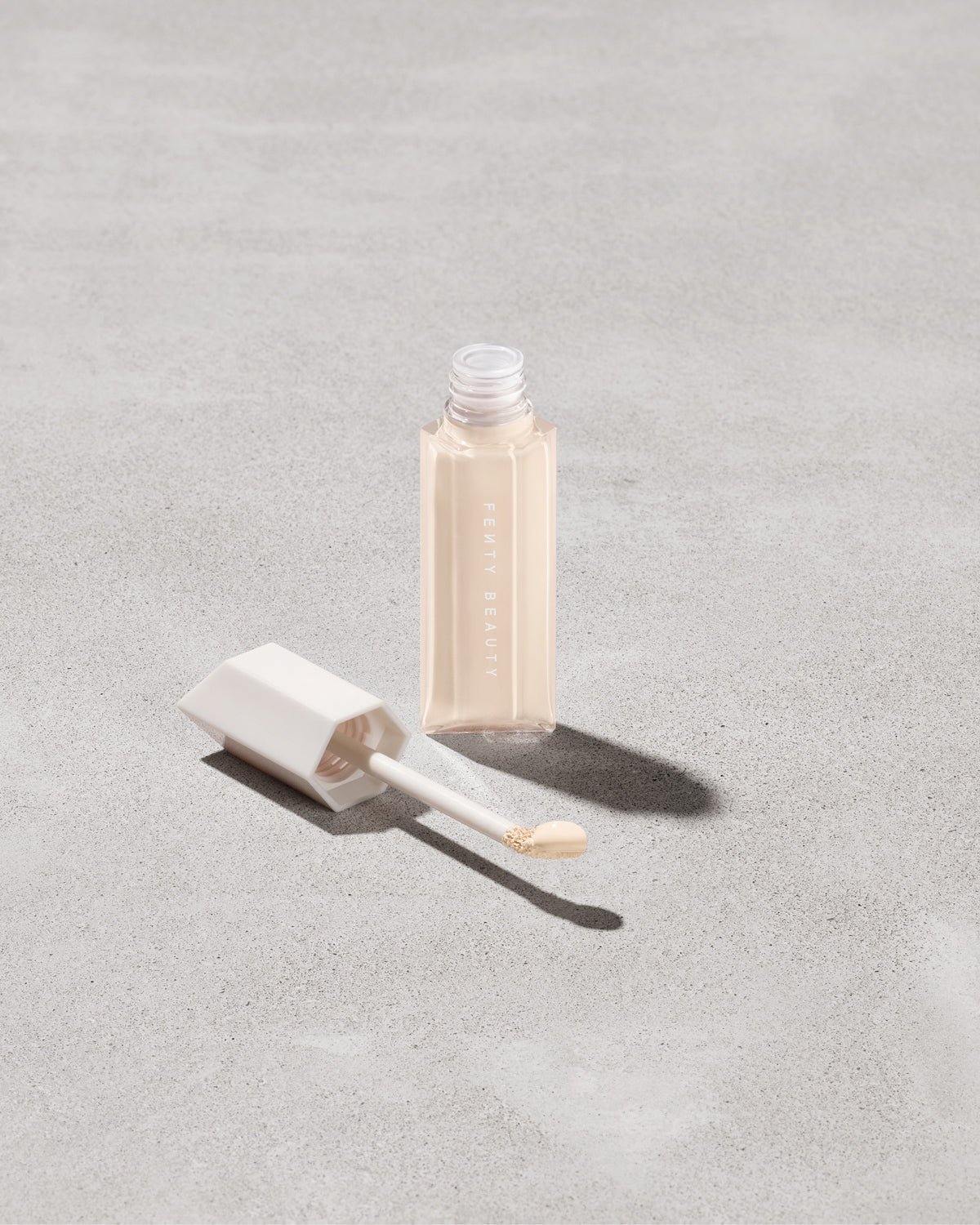 FB_SPR23_T2PRODUCT_ECOMM_WERE_EVEN_110W_sRGB72 Open bottle of Fenty Beauty We're Even Hydrating Longwear Concealer with doe foot applicator in shade 110w on a concrete background.