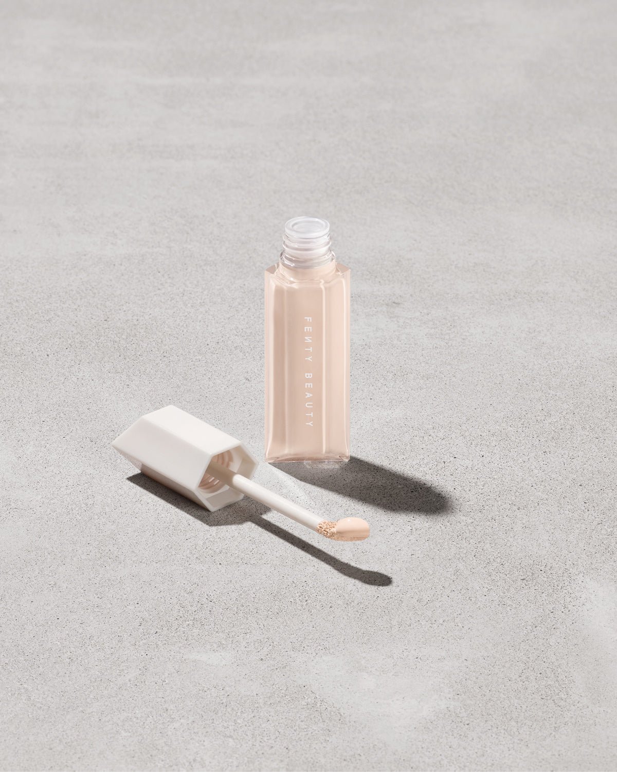 FB_SPR23_T2PRODUCT_ECOMM_WERE_EVEN_125C_sRGB72 Open bottle of Fenty Beauty We're Even Hydrating Longwear Concealer with doe foot applicator in shade 125c on a concrete background.